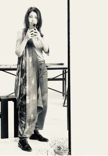 Darshana Shilpi, founder of Alba Amicorum, taking a mirror selfie wearing a long coat and jeans indoors.
