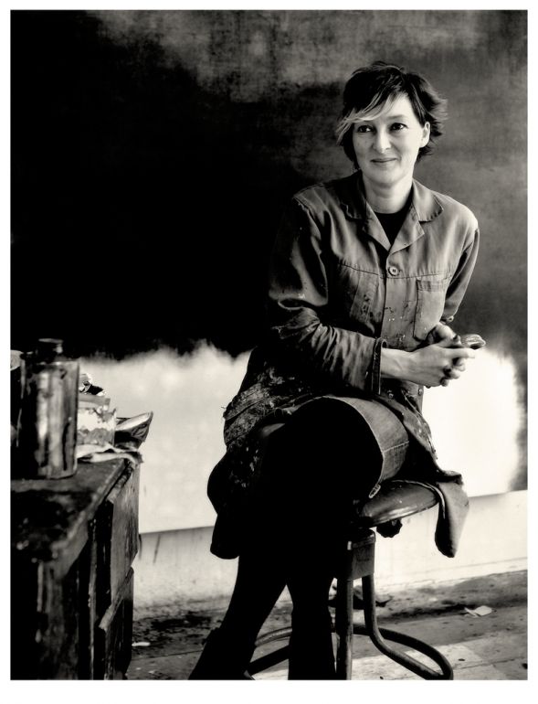 Black and white photo of artist Naomi Frears sitting on a stool with a large painting behind them