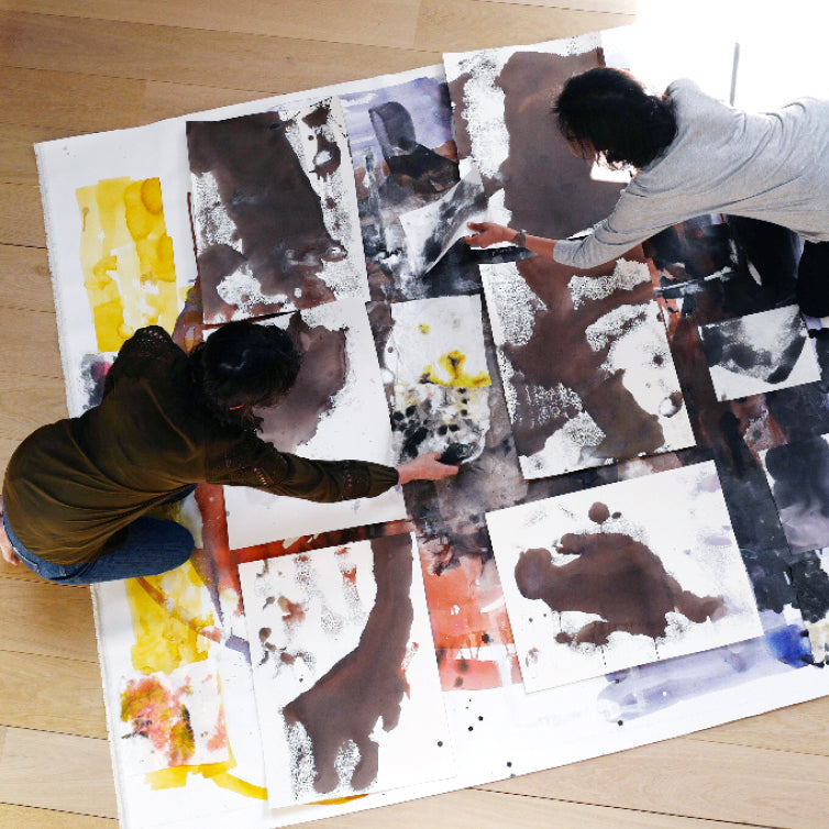 two artists kneeling over a large canvas collaborating on a painting & collage