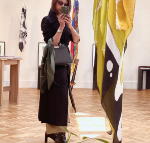 A woman photographing herself in the mirror, phone partially blocking her face - she is wearing an art scarf of a Man Ray Ray-o-graph, sunglasses, a long coat, and black boots and standing next to a gently hanging large silk scarf