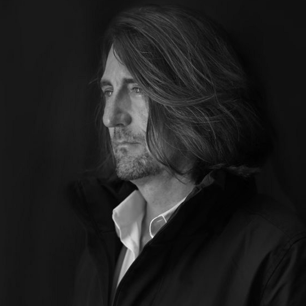 black and white profile photo of Jorg Brauer in a dark jacket and white collared shirt, his long hair is swept back