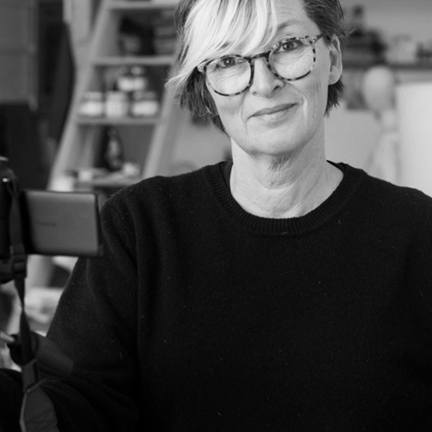 photo of artist Naomi Frears wearing glasses and a black sweater, smiling in her studio