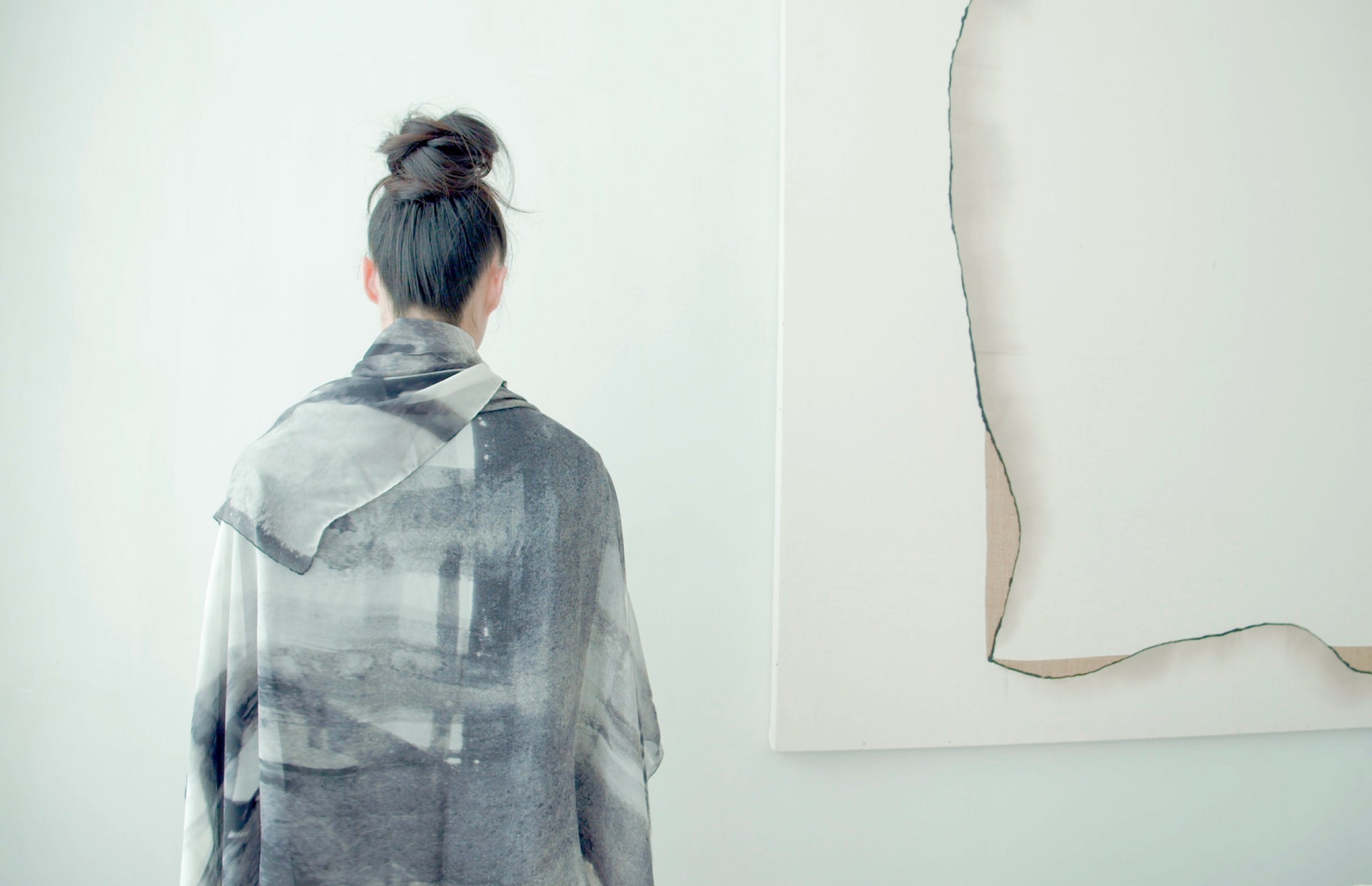 A woman draped with Autumns Rendition abstract printed scarf in pale greys standing before a mostly white painting