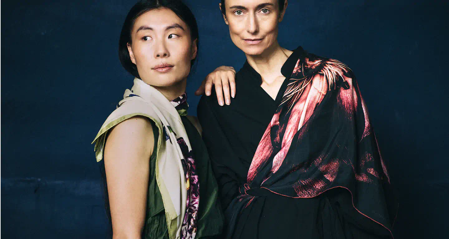 Two women wearing colorful silk Alba Amicorum scarves by James T Murray and Darshana Shilpi Rouget against a dark backgroun
