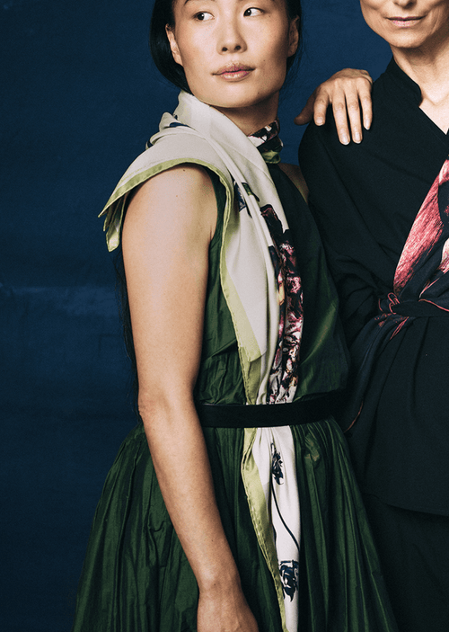 Two women standing close together against a dark blue background, wearing patterned scarves.