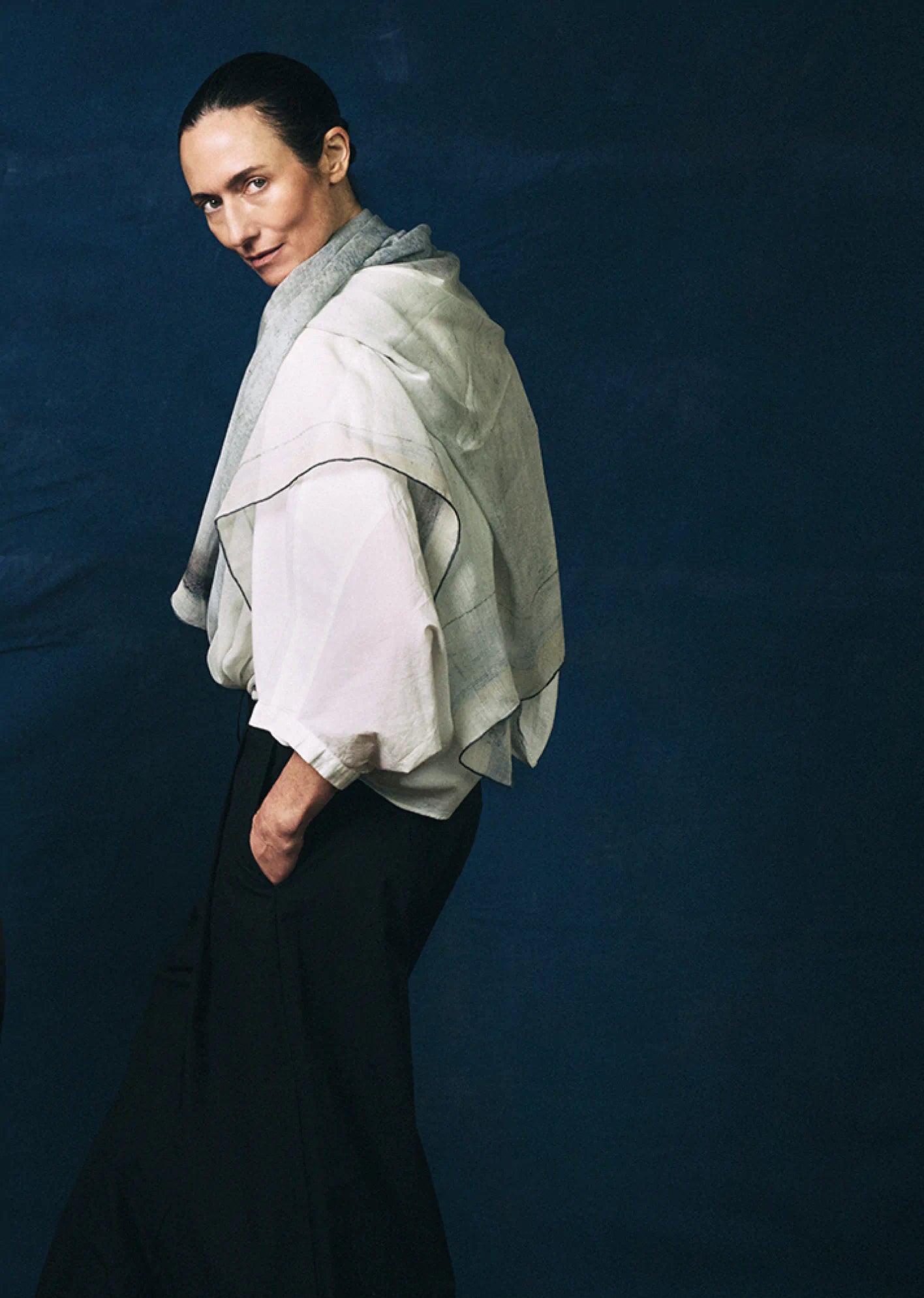 Woman in a white shirt and dark pants with a Dot Wade scarf wrapped around her waist and flowing behind her, standing against a dark blue background