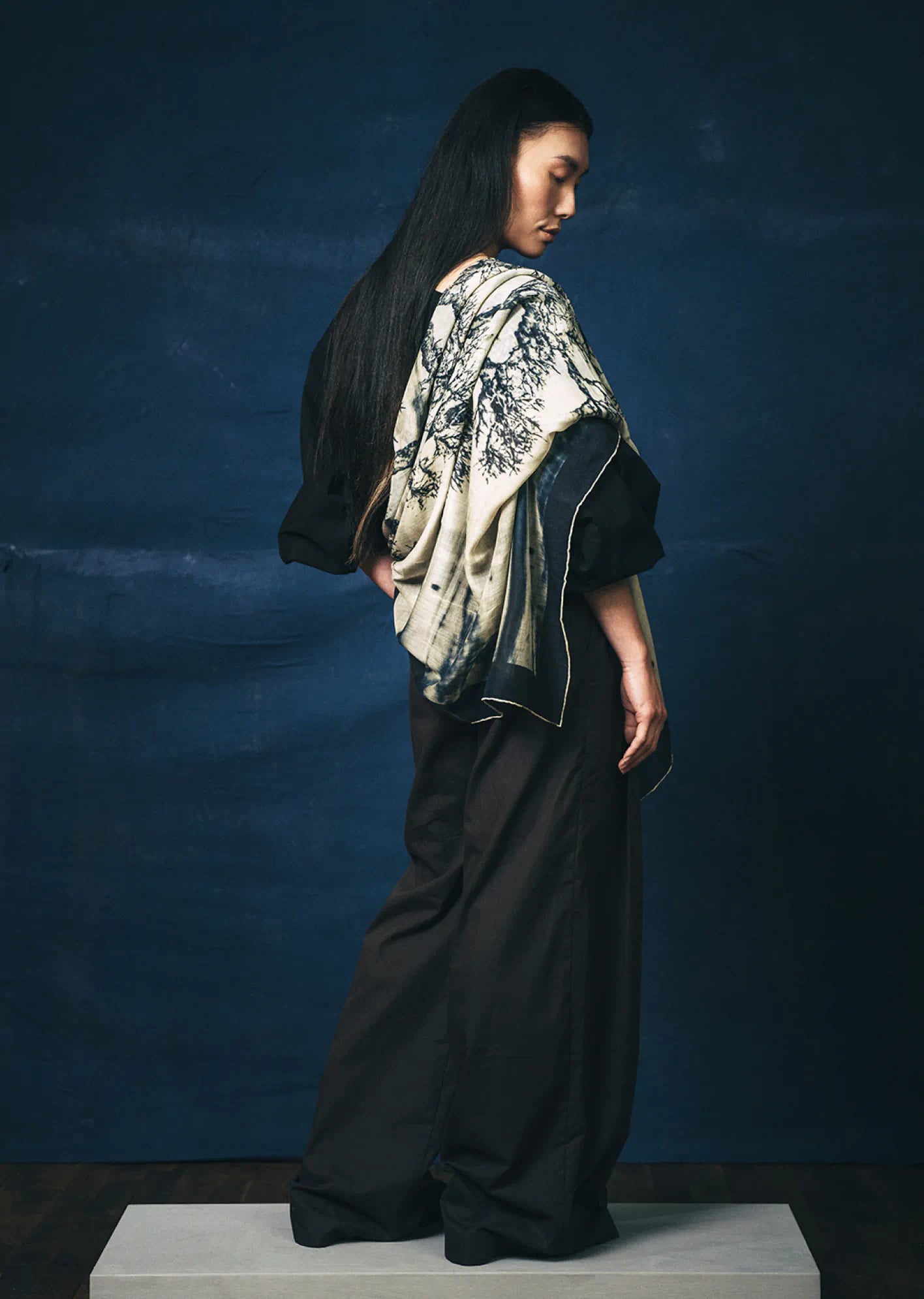 Person with her back to the camera gazing down, wearing a black outfit with a James T Murray patterned scarf against a dark blue background