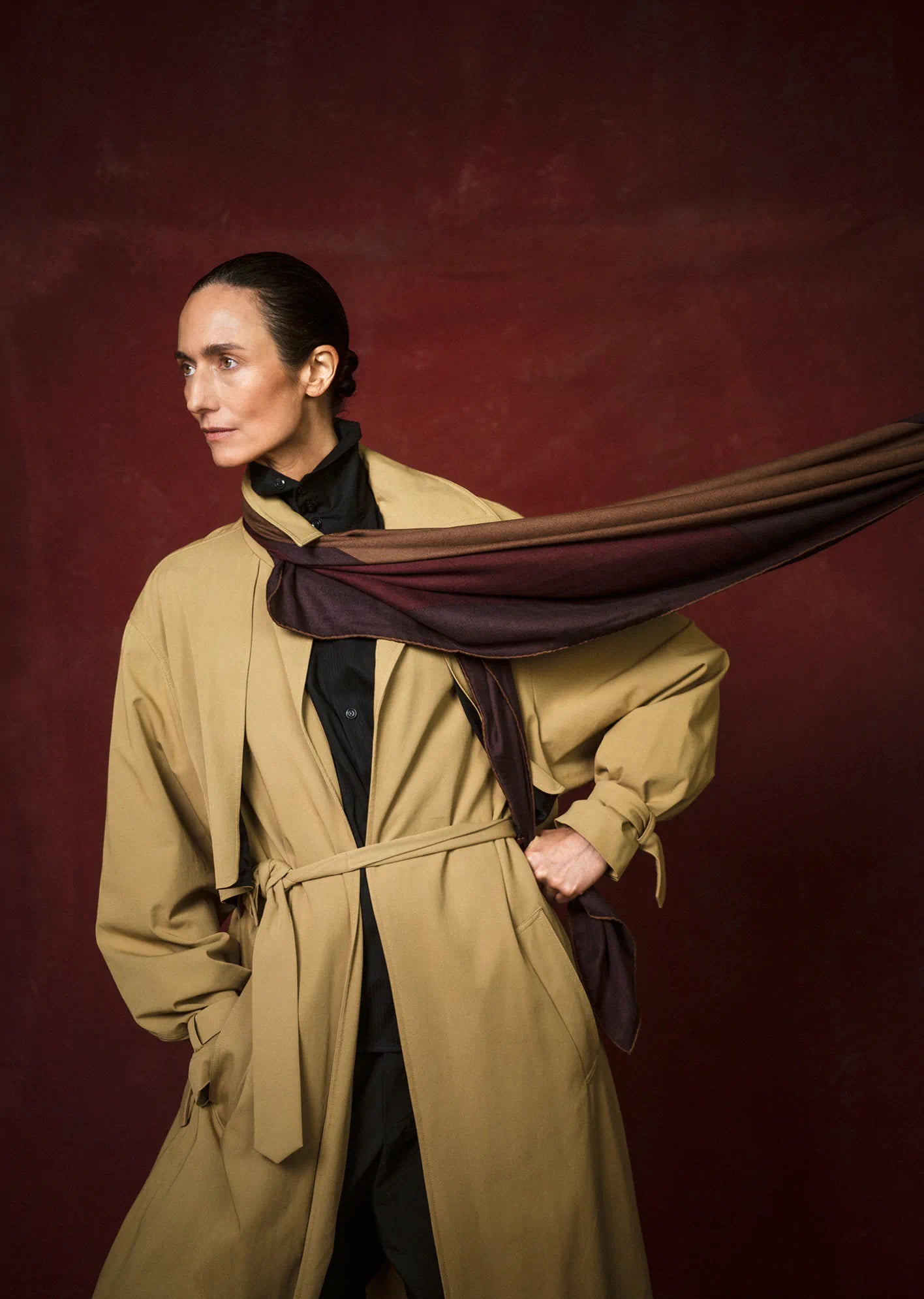 Woman wearing a beige trench coat with a dark scarf against a red background