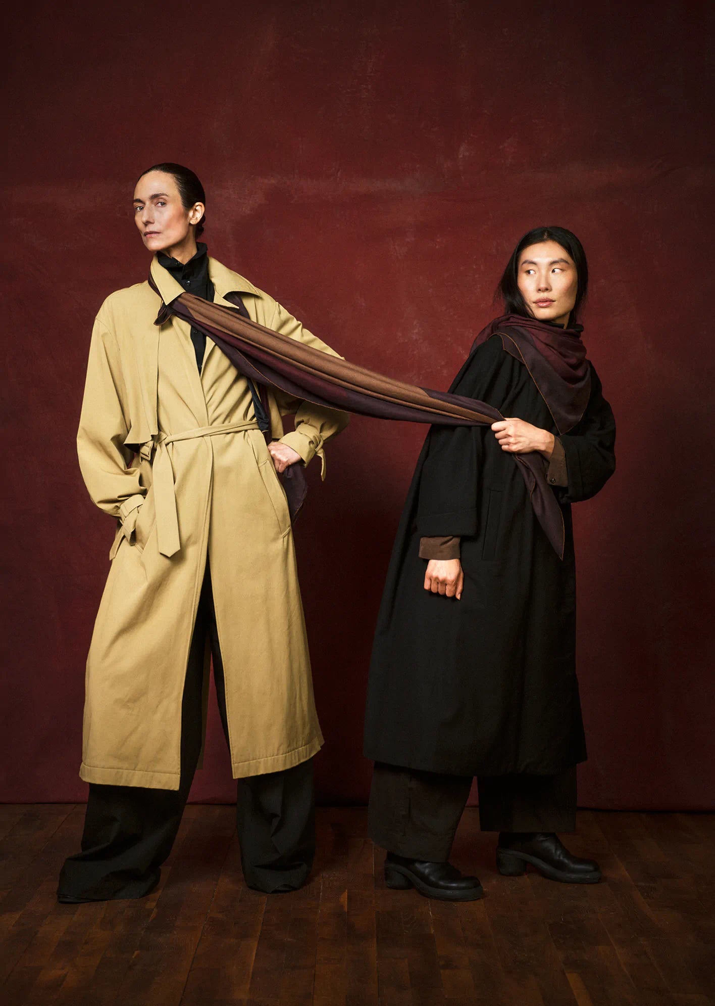 Two people wearing trench coats and scarves by Darshana Shilpi Rouget against a dark red background