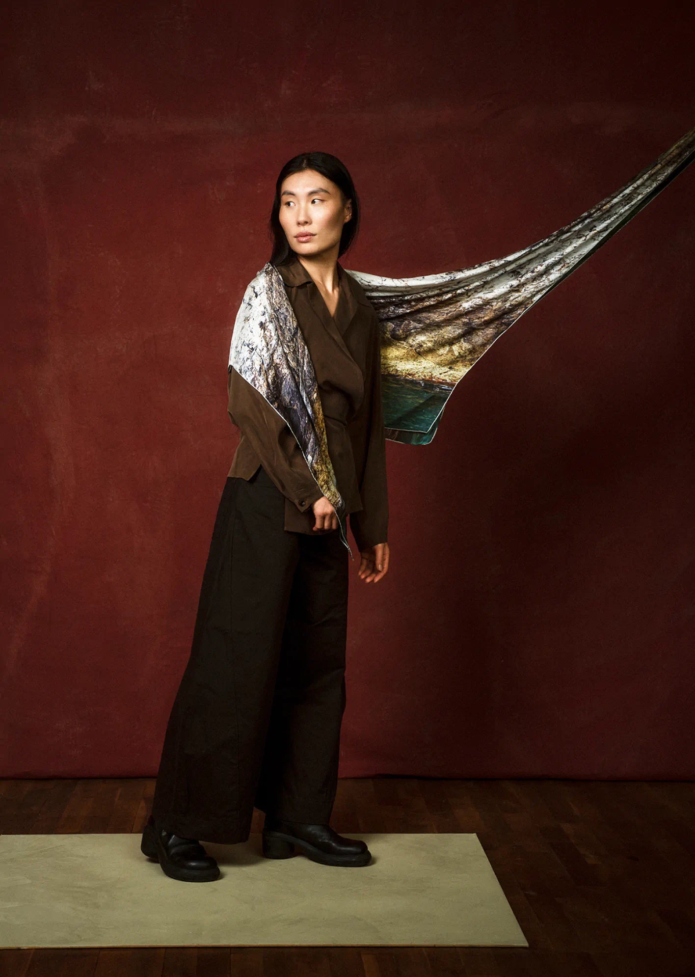 Person holding a large art scarf by Jorg Brauer with a scenic design against a dark red wall