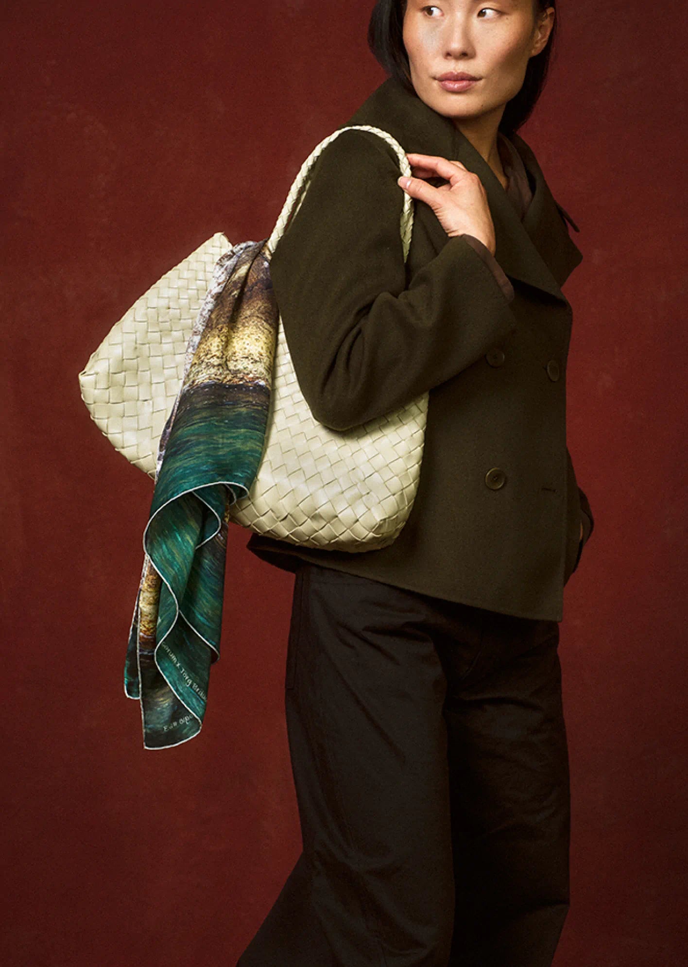 Person holding a large textured bag draped with an art scarf by Jorg Brauer with a scenic design against a dark red wall