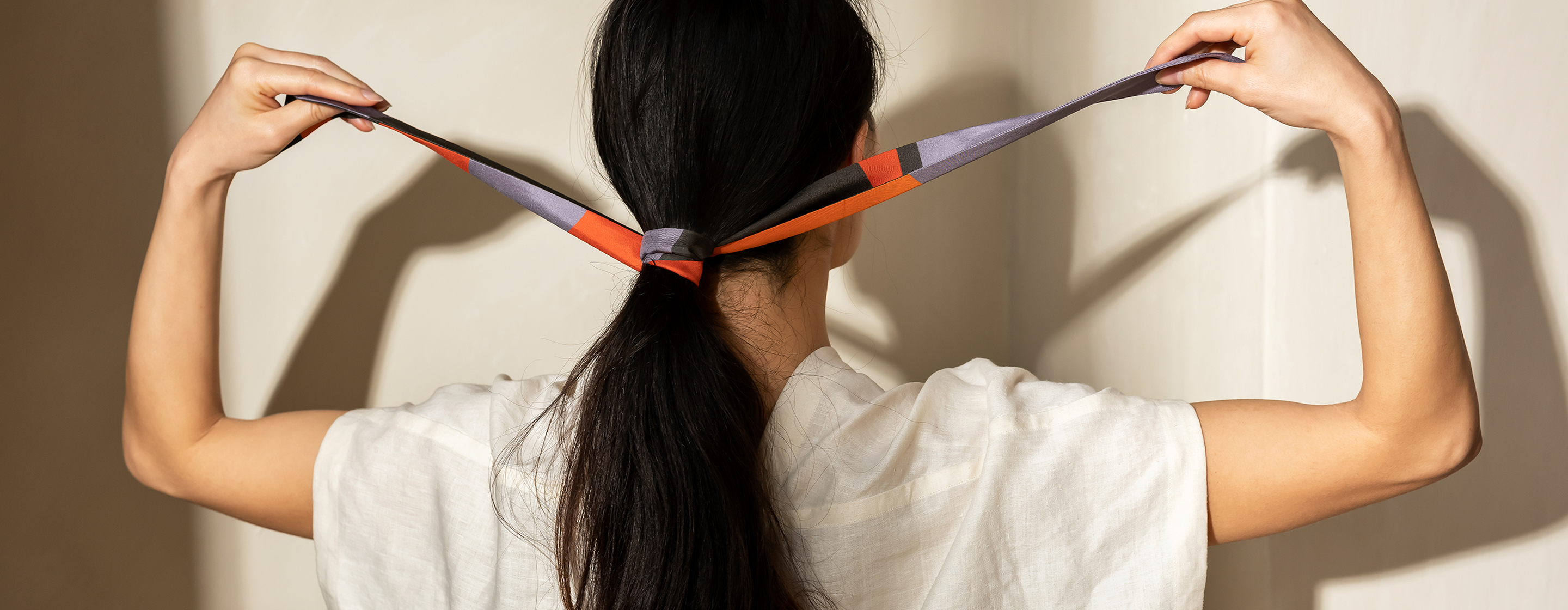 Person seen from the back tying an Alba Amicorum Twilly around thieer long dark hair against a neutral background