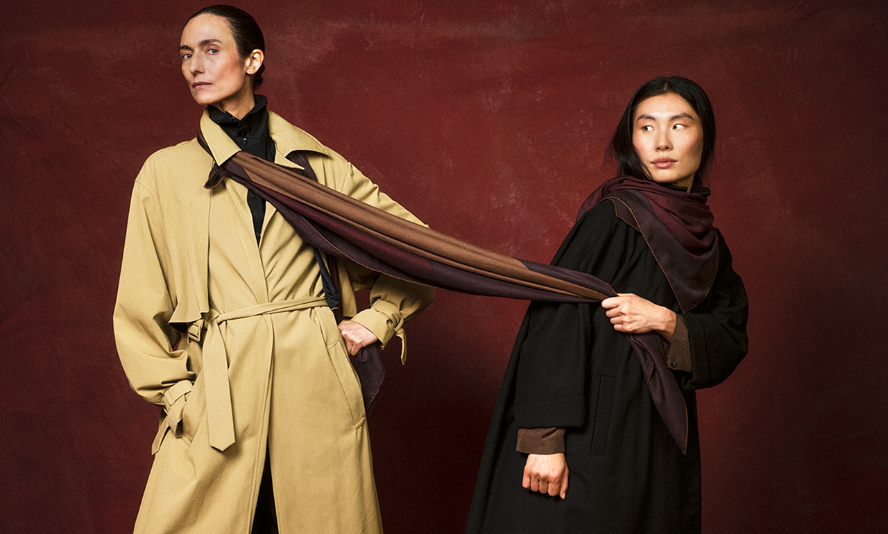 Two women holding a long brown scarf against a dark red background