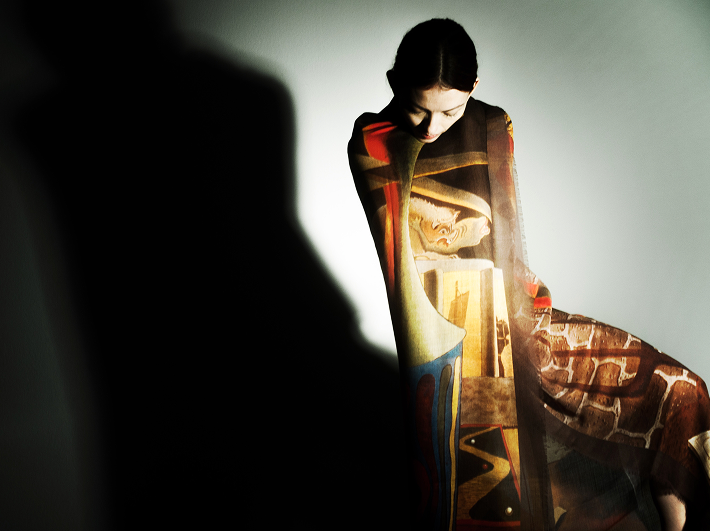 Person wrapped in a colorful shawl with an image from the artist, Man Ray, on it, seated against a plain background.