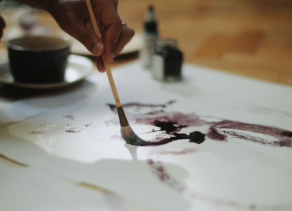 Person painting with a brush on a white surface, with ink and a cup in the background.