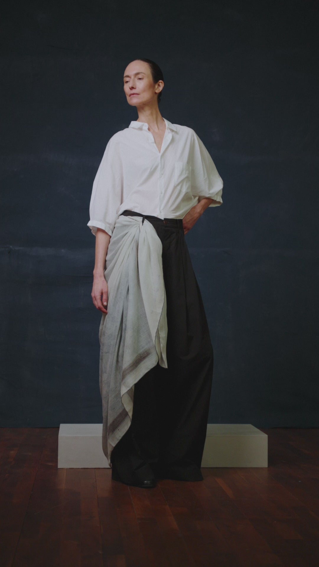 VIDEO OF A Woman in a white shirt and dark pants with a Dot Wade scarf wrapped around her waist and flowing behind her, standing against a dark blue background