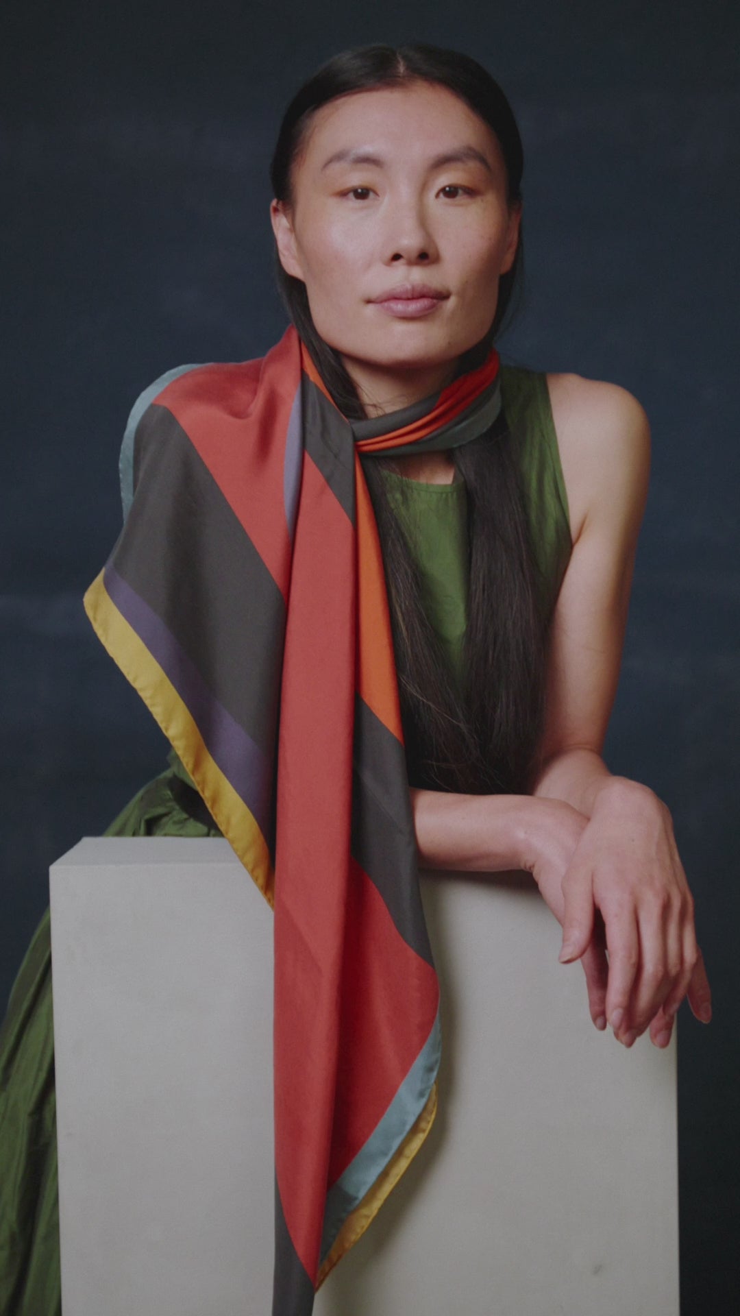 video of a woman standing behind a pedestal wearing a colorful Alba Amicorum silk scarf in front of a dark background 
